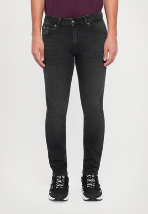 Jeans negros ajustados con una textura suave, con bolsillos delanteros y acentos de metal. Llevados con zapatillas negras que tienen letras blancas.