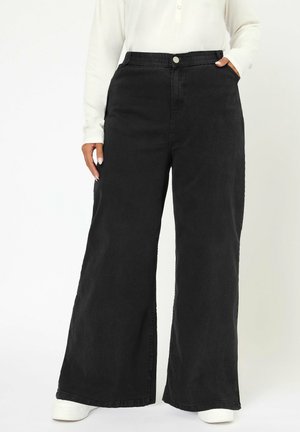 Person trägt schwarze High-Waist Jeans mit weitem Bein, elastischem Bund und Knopf, kombiniert mit weißen Schuhen und einem weißen Langarmshirt.