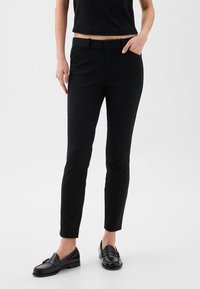 Femme portant un pantalon noir slim, des mocassins noirs et un top court noir, debout avec une main dans la poche devant un fond uni.