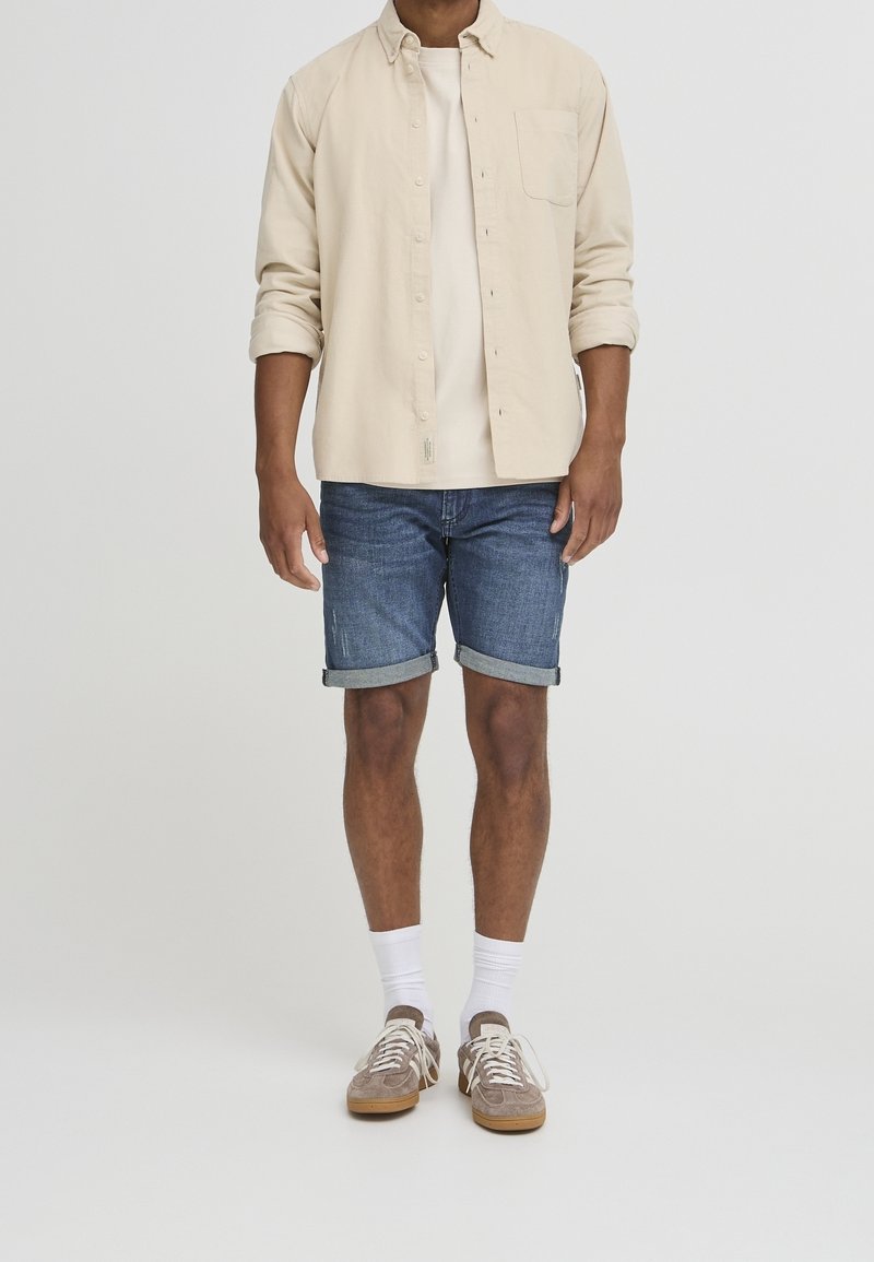 Camicia beige a maniche lunghe con bottoni sopra una t-shirt chiara, abbinata a pantaloncini in denim e sneaker beige con lacci bianchi e calzini.