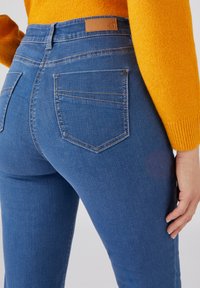 Jean en denim bleu à coupe ajustée, dotés de deux poches arrière avec des détails de couture et d'un patch en cuir marron sur la taille.