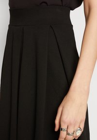 Černá plisovaná sukně s širokým pasem a hladkým, texturovaným materiálem. Ruka se spoustou stříbrných prstenů leží na sukni.