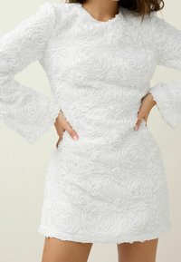 Robe mini blanche en tissu texturé avec des motifs en forme de roses en 3D, des manches longues en forme de cloche et un col rond.