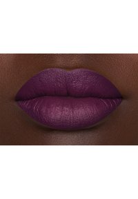 Rossetto opaco di colore viola profondo applicato su labbra piene, che mettono in mostra una texture liscia e una forma definita contro uno sfondo di tonalità di pelle più scura.