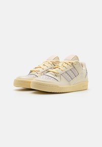 Beige leather sneakers with pastel purple stripes, perforated toe, and yellow laces. Features a textured rubber sole and retro design.