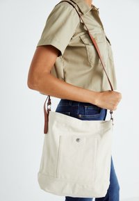 Borsa tote in tela color crema con una tracolla in pelle marrone, dotata di una tasca frontale e una chiusura a scatto. Design semplice con materiale resistente.