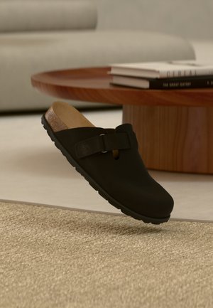 Μαύρο παπούτσι slip-on με φελλό πάτο και ρυθμιζόμενο λουράκι, αιωρείται πάνω από μπεζ υφασμένο χαλί, με ξύλινο τραπεζάκι καφέ και βιβλία στο φόντο.