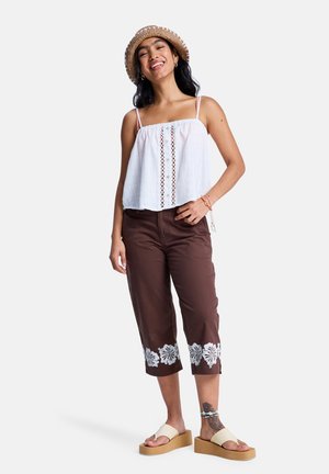 Weißes Baumwoll-Crop-Top mit Spitzendetails, kombiniert mit braunen, kurz geschnittenen Hosen mit weißer Blumenstickerei am Saum und braunen Plateau-Sandalen.