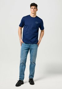 T-paita, tummansininen, puuvillaa, pyöreä kaula-aukko ja lyhyet hihat, jossa on pieni grafiikka rinnassa. Yhdistetty vaaleansinisiin farkkuihin ja mustiin kenkiin.