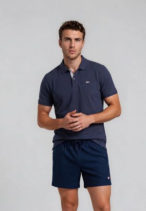 Joven de pie con camisa polo azul marino y pantalones cortos a juego, posando frente a un fondo liso gris claro.