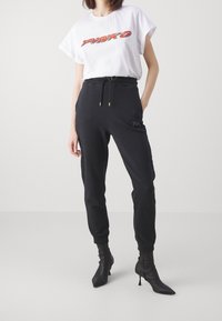 Bílý bavlněný tričko s červeným logem, spárované s černými tepláky s kapsami a stahovací šňůrkou, doplněné černými špičatými kotníkovými botami.