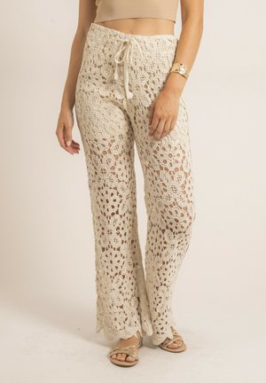 Femme portant un pantalon en dentelle crochet beige à motifs floraux, taille nouée, bracelet en or et sandales dorées, debout devant un fond uni.