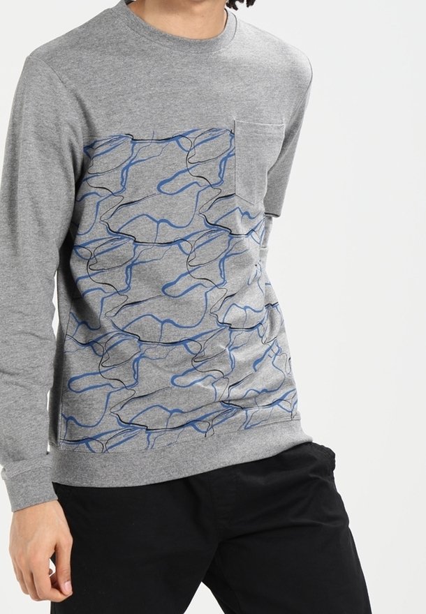 Homme portant une chemise grise à manches longues avec des motifs abstraits de lignes bleues et noires et une poche poitrine, associée à un pantalon noir.