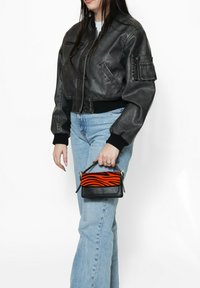 Giacca bomber nera in pelle, taglio corto, con polsini a costine; modello che tiene in mano una piccola borsa a strisce nere e arancioni. Jeans in denim blu chiaro.
