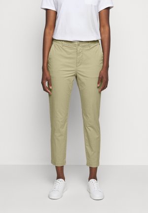 Osoba nosząca beżowe, przycięte spodnie, białą koszulę z krótkim rękawem włożoną w spodnie oraz białe tenisówki na sznurówki, stojąca na tle jednolitego jasnego tła.