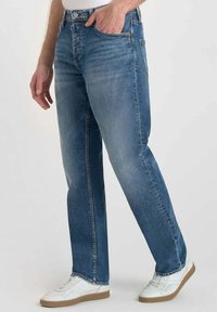 Jean en denim bleu délavé avec une coupe droite, un design à cinq poches et des coutures décoratives. Associés à des baskets blanches.