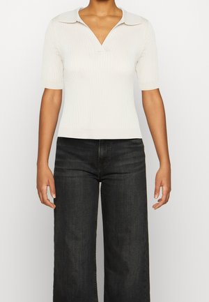 Kobieta ubrana w biały, prążkowany sweter z krótkim rękawem, szerokim kołnierzem i czarne jeansy z szerokimi nogawkami, stojąca na jednolitym tle.