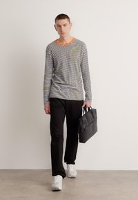 Camicia a maniche lunghe a righe con scollo arancione, pantaloni neri e sneakers bianche. Il modello porta una borsa tote nera, su uno sfondo chiaro e semplice.