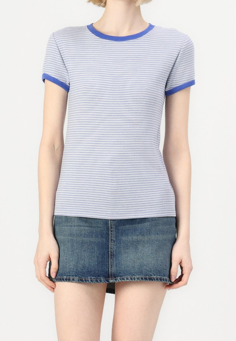 Femme portant un t-shirt à manches courtes rayé bleu et blanc avec des bordures bleues et une mini-jupe en jean délavée, debout devant un fond uni.