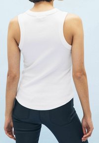 Débardeur blanc à côtes avec un col rond et de grandes emmanchures, présenté avec un ourlet arrondi et associé à un pantalon noir ajusté.