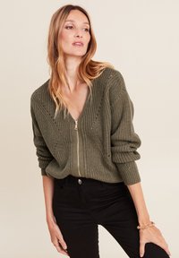 Olivgrüner Reißverschluss-Pullover mit strukturiertem Strickmuster, V-Ausschnitt und tiefen Schultern. Kombiniert mit schwarzen Hosen und einem Armband.