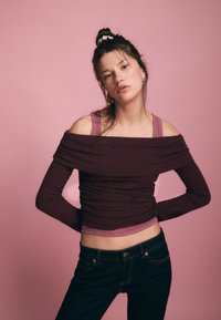 Schulterfreies, bordeauxrotes Ripp-Top mit langen Ärmeln und figurbetontem Schnitt, darüber ein hellviolettes Tanktop. Dunkle Jeans aus Denim vervollständigen das Outfit.