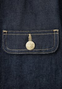 Tissu en denim bleu foncé avec un petit rabat de poche rectangulaire sécurisé par un seul bouton métallique décoratif.