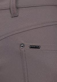 Grijze stoffen broek met gestikte naden, een zijzak en een metalen knoopaccent. Bevat een merketiket op de zak.