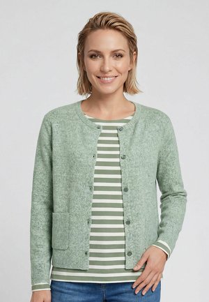 Woman smiling wearing a light green buttoned cardigan over a green and white striped long-sleeve shirt and blue jeans against a plain background.