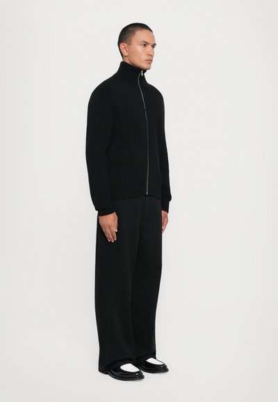 Black ribbed zip-up sweater with high collar, paired with wide black trousers and black shoes featuring white accents. Front view.