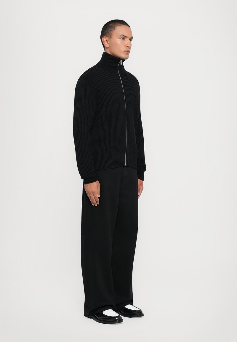 Pull en maille noire zippé avec col montant, associé à un pantalon large noir et des chaussures noires avec des accents blancs. Vue de face.