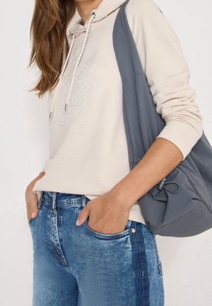Woman with long brown hair wears a beige hoodie, blue jeans, and a gray shoulder bag, with one hand in her pocket.