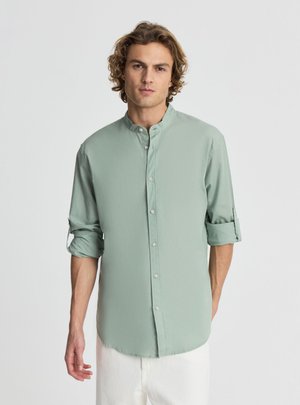 Giovane uomo con capelli ricci che indossa una camicia verde chiaro con collo mandarino e maniche risvoltate e pantaloni bianchi, in piedi davanti a uno sfondo semplice.