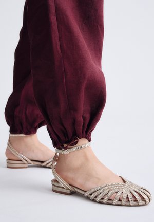 Pies con sandalias planas beige con tiras y pequeñas cuentas, combinadas con pantalones burdeos con puños ajustables, sobre un fondo neutro.