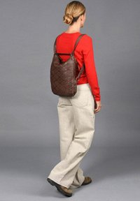 Bruine leren rugzak met een gequilt patroon, verstelbare banden, gedragen met een rode top en beige wijde broek.