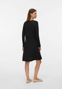 Robe noire à manches longues avec un design de top court, tissu texturé, jupe évasée et coupe mi-longue ; associée à des ballerines de couleur claire.