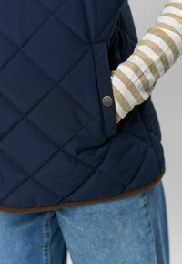 Námornícka prešívaná vesta s hnedým lemom, s bočnou kapsou na kovový cvok a pruhovanou košeľou s dlhým rukávom pod ňou.