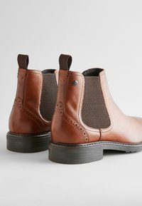 Brown leather Chelsea boots with elastic side panels, decorative brogue detailing, and a black rubber sole. Pull tabs at the back for ease of wear.
