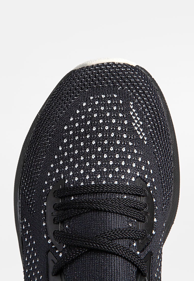 Zapatilla deportiva negra con parte superior de malla texturizada, que presenta perforaciones blancas y cordones negros. La zona del dedo tiene una forma redondeada.
