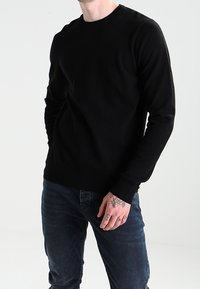 Zwarte langemouwen trui met ronde hals, gemaakt van zachte stof, gedragen over blauwe jeans. Heeft aansluitende mouwen en een gladde textuur.