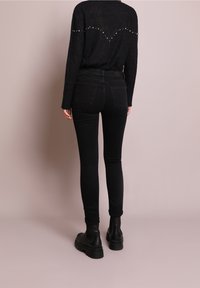 Czarny dzianinowy sweter z gwiaździstym wzorem w kształcie litery V na plecach, zestawiony z obcisłymi czarnymi dżinsami i czarnymi botkami na płaskim tle.