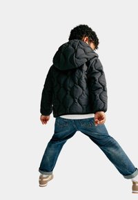 Giubbotto nero trapuntato con cappuccio, abbinato a jeans blu con risvolti e sneaker marroni, visto da dietro.