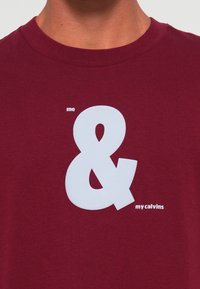 Bordo krūšu tīkla krekls ar lielu baltu ampersandu grafiku un mazu tekstu, kas lasāms "me" virs un "my calvins" zem krūtīm.