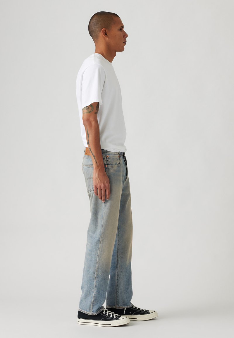 Weißes Baumwoll-T-Shirt, lockere Passform, kombiniert mit hellblauen Jeans, leicht ausgewaschen. Schwarze Leinensneaker mit weißen Sohlen. Seitenansicht.