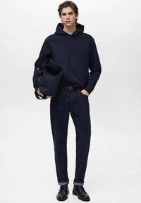 Marinblå hoodie, mörka jeans i denim, svarta skor. Modellen håller en mörk väska. Enkel design, standardpassform, minimal detaljering.