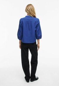 Camicia blu a maniche corte con dettaglio sul retro, abbinata a pantaloni neri a gamba dritta e scarpe nere. Stile semplice e casual.