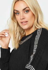 Svart ribbad tröja med silverdetaljer längs axlarna och halsringningen. Har lång ärm och en strukturerad yta.