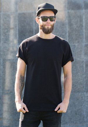 Black short-sleeve t-shirt, relaxed fit, with rolled cuffs. Paired with black cap and sunglasses. Skin visible on arms with tattoos.