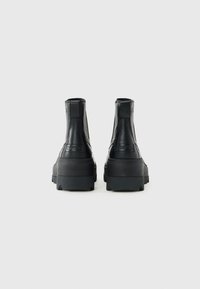 Sorte lær ankle boot med tykke gummisåler, sett fra baksiden, på en enkel lys grå bakgrunn.