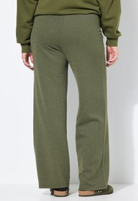 Persoon in een textuur met olijfgroene wijduitlopende broek, bijpassend lang mouwen top en donkere instap schoenen, staand tegen een witte muur.